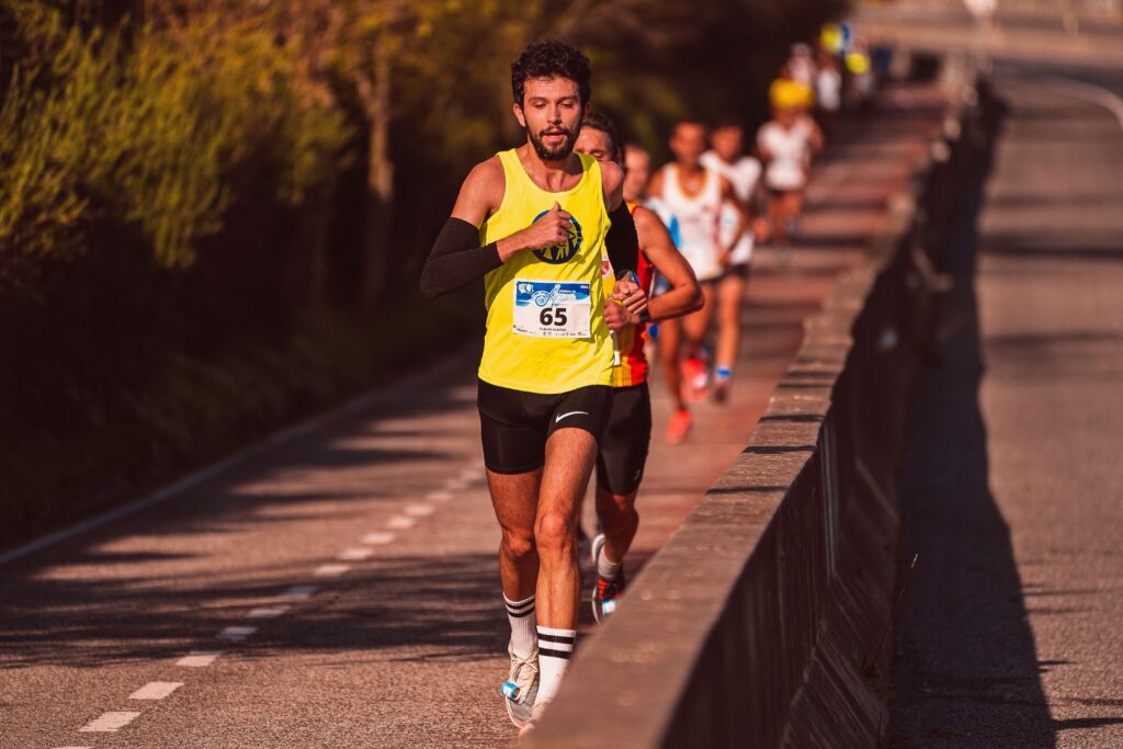 pexels-photo-10417359-10417359 Focused male athlete running in a marathon on a sunlit road, leading a group of runners.